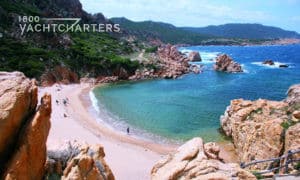 Photograph of a secluded cove in Sardinia. View from the top of a rocky cliff, looking down into a cove with turquoise water and beach.