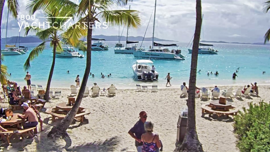 Photo of beach with people sitting in beach chairs. There are multiple sailboats anchored in the water just off of the beach. Palm trees sway on the beach.,