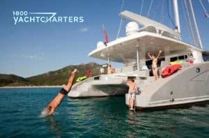 Photograph of a catamaran sailboat at anchor, while charter guests sit with feet in the water or dive off and swim from the boat.