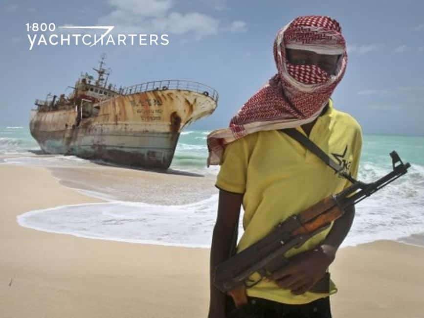pirate. Photograph of a soldier carrying a rifle on a beach in front of a decrepid ship. The soldier is wearing a yellow t-shirt and red shawl around his head. You can see his dark skin on his arms and his eyes. The boat behind him is just the shell of a large ship.