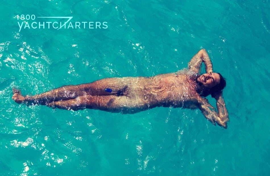 Photo of a man floating on his back in turquoise water. He is nude. His genitals are blurred out of the photo. He has tan lines where his shorts would be. 