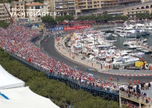 Aerial photograph of monaco grand prix race track. To the left of the photo are people in the bleachers, track in the middle (no cars on it in picture), and to the right are large yachts docked next to the race track, behind barricades