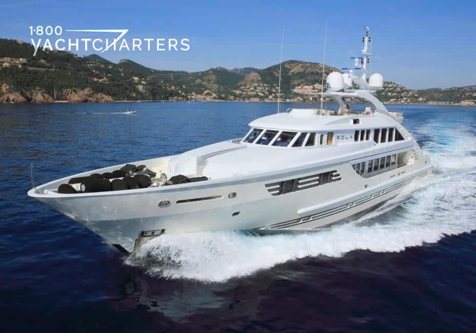 Photograph of motoryacht OLA running. Yacht is heading to the left bottom corner of the screen. There are rocky hillsides in the background, as well as a tiny yacht anchored in the background.