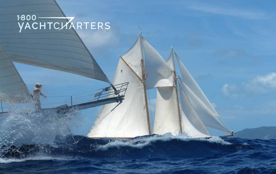 Classic yachts. Photograph of two schooner sailboats underway. The one in the foreground only shows the bowsprit and part of the front sail. The yacht in the background is fully under sail, and a large wave is lapping at its side.