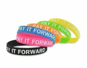 Photograph of Pay It Forward bracelets in a stack. There are 6 bracelets, and they are all different colored. All have white text that says, Pay It Forward, in all caps. Silicone bracelets.