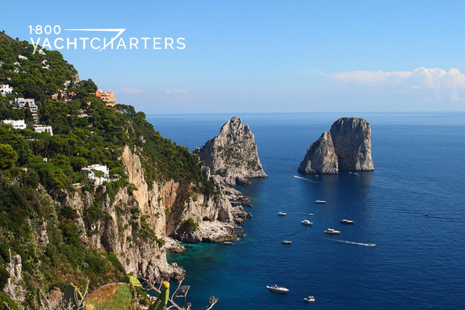 Aerial photograph of Capri, Italy.  Photo shows a few boats in the bay, one of them in motion.  It also shows a cliff with a few homes dotted on it on the left side of the photo, and 2 huge boulder rocks jutting up from the sea in the center and right side of the back of the photo.  Deep blue water.