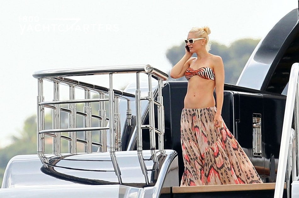 Photograph of Gwen Stefani talking on the telephone while standing at the back of a yacht. She is wearing a long flowing skirt and a bikini top. Her blonde hair is up on top of her head, and she is wearing white sunglasses