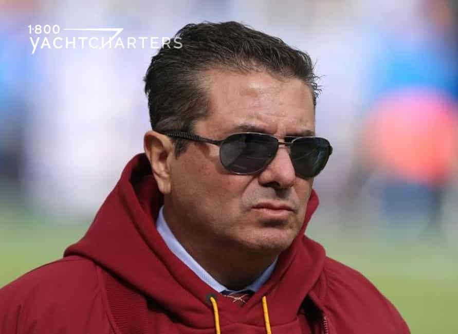 Headshot photograph of Dan Snyder, owner of NFL team, Washington Redskins. He is wearing a red jacket and dark sunglasses. He has a scowl on his face.
