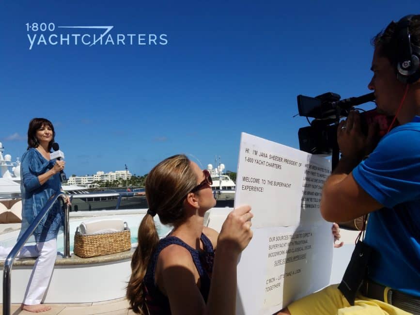 Photograph of a girl with microphone being filmed. She is speaking on the top deck of a yacht. A film crew holding cue cards stands in front of her, out of camera view.