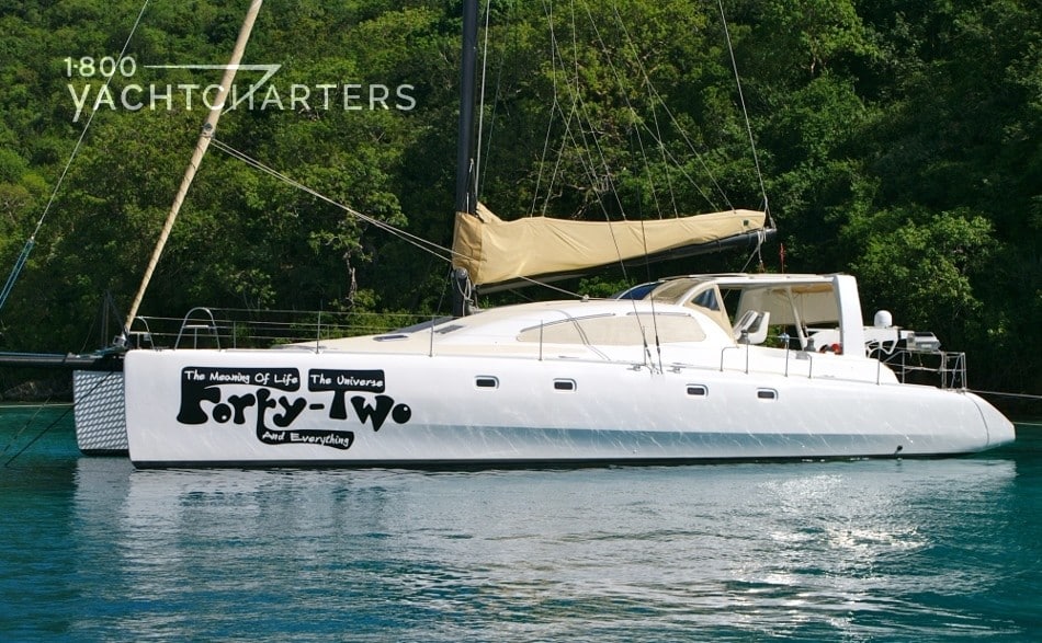 Profile photograph of a catamaran sailboat at anchor. The sails are down and wrapped in beige sail coverings on the mast. The sailboat is white with thick black font writing garbled at front of yacht. The yacht is facing the left side of the picture