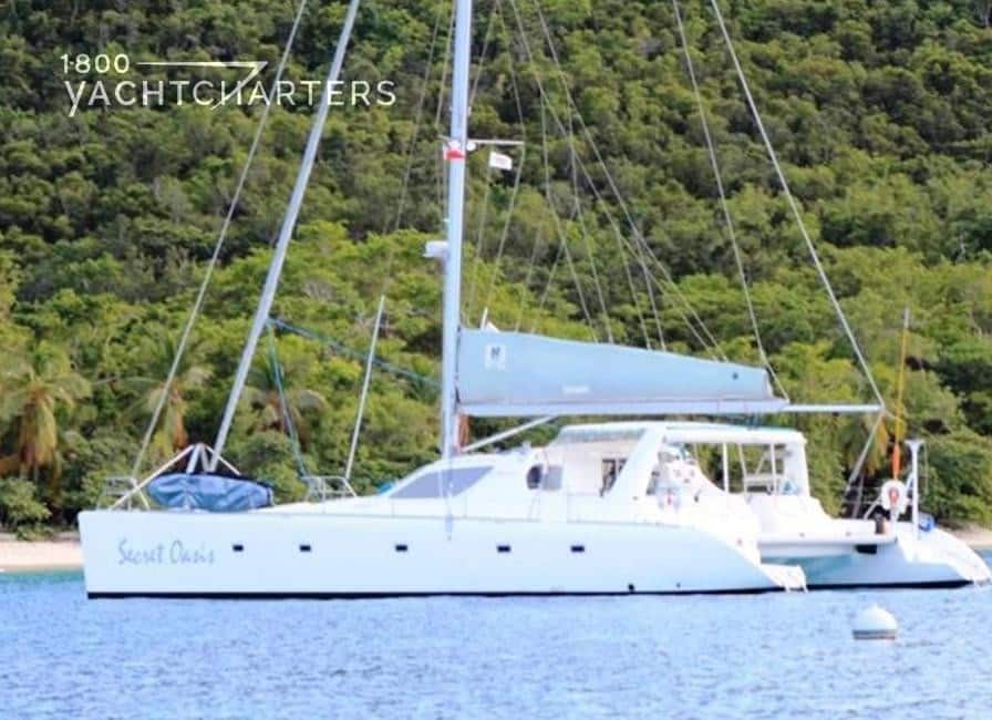 Profile photograph of sailing catamaran, Secret Oasis. The name is on the side of the boat in blue cursive font. The yacht is at anchor. Her sails are down. There is a light blue sail cover over the sail that goes horizontally across the boat.