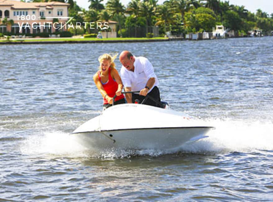Photograph of a man and woman riding on a white circraft (a round watertoy). In the background, on the left side of the photo, is a huge mansion on a waterway.