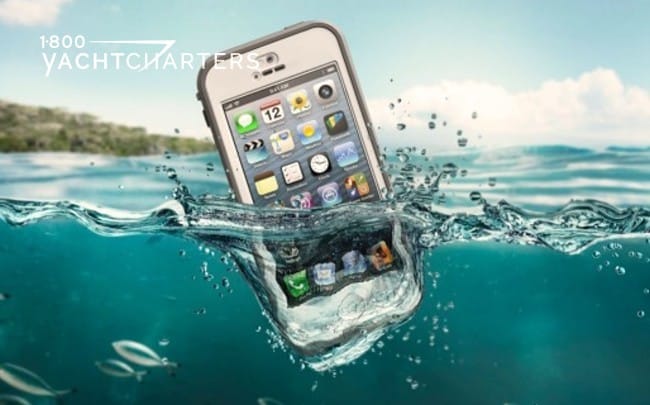 Photograph of a cellphone that has just fallen into the ocean. There is a splash wave on the right side of it. It is a white phone with apps visible on the front. There is an island in the background of the photo..