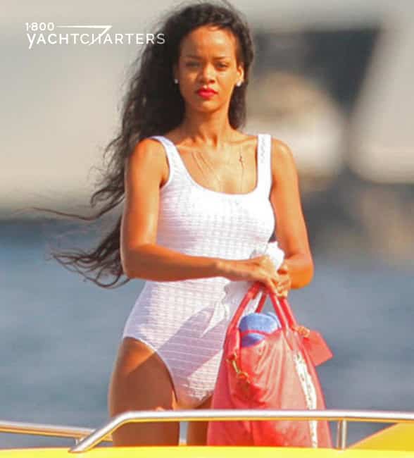 Photograph of celebrity, Rihanna, in a white one-piece swimsuit on a yellow powerboat. There is a large white yacht in the background (blurred out).