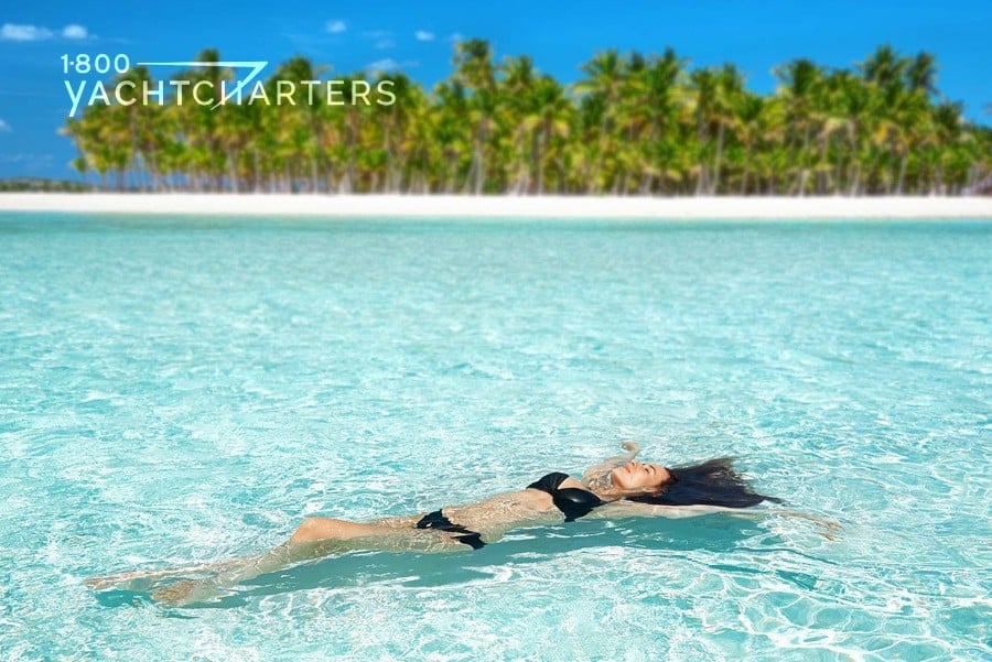 Photograph of a woman in a black bikini who is floating in the water off of a beach. The beach is clear turquoise. The sand in the background is bright white. The trees are lush and green. Tropical photo. 