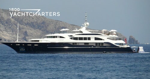 Profile photograph of motoryacht Swan by Benetti. She is facing the center left side of the photo. There is a large rock mountain in the background. The yacht hull is dark, and the superstructure is white.