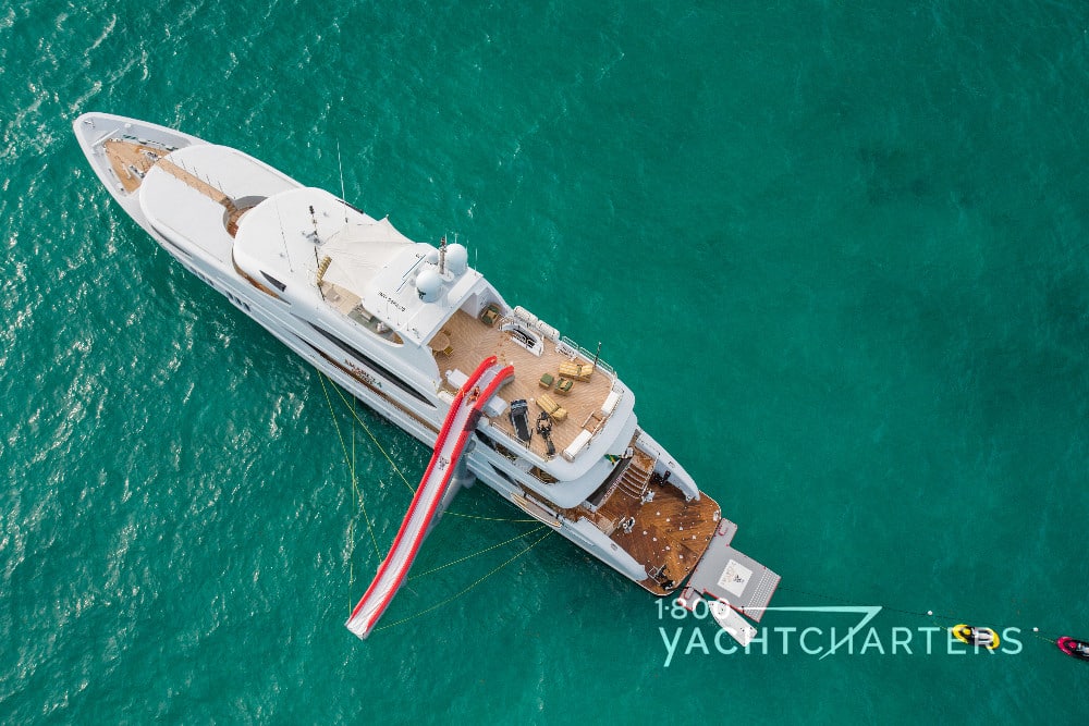 Aerial view of inflatable red and white slide deployed from the top deck of the yacht, Amarula Sun. Jet skis tied behind the boat. The vessel is at anchor in calm, green waters. 