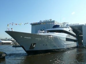 Photograph of a large white superyacht at launch. She is anchored and has colorful banner flags having across the length of the ship. There is a large building behind her