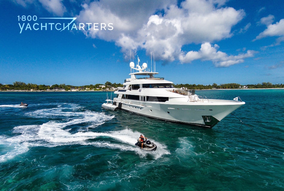 Photograph of motoryacht W at anchor. The yacht is facing the right side of the photo. Someone is riding a jetski next to the yacht, headed to the right, also