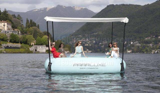 Photograph of a floating sea cabana. There are 3 people on it. There are mountains in the background.