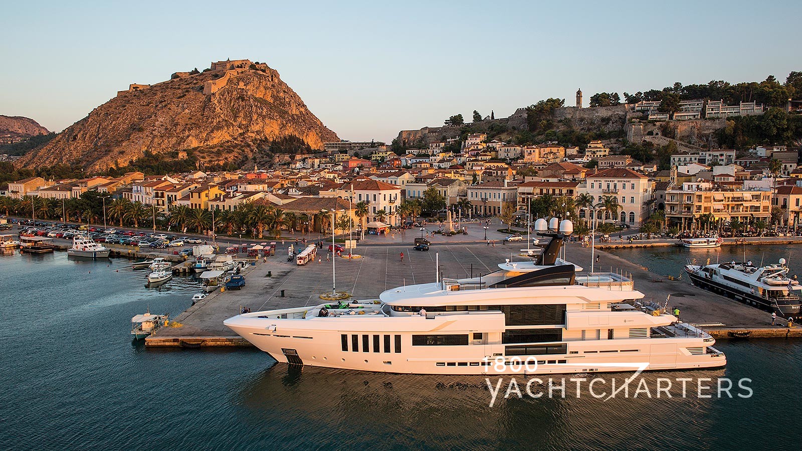 Profile photograph of a white yacht anchored in a marina.  She is the largest yacht in the marina. There are many small boats behind her.  There are mountains in the background.  The sun is setting in front of the yacht, so she is bathed in golden light.