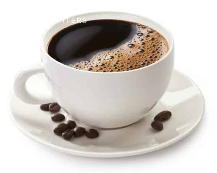 Photo of a cup of coffee with white cup and saucer. There are 10 coffee beans spread across the white saucer.  There are caramel-colored bubbles across one half of the liquid in the coffee cup.  The other half looks dark brown.