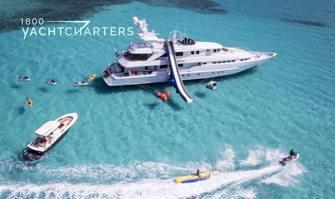 Aerial photo of yacht at anchor with an inflatable slide coming down from the top deck. Multiple jetskis and tenders are racing around in the water around the boat. The water is clear and turquoise.