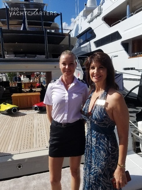 Jana Sheeder, President of 1-800 Yacht Charters standing with yacht Vertige female crewmember on dock in front of boat. Jana wearing halter long dress and gold name badge, and crewmember wearing short black skirt and white short-sleeved shirt.