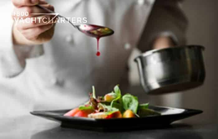 Photo of a chef in white clothing standing over a black shiny table. He has a silver spoon over a plate of food. There is red juice dripping off of the spoon down onto the plate. The chef is Tankoa yacht Solo gourmet chef. 