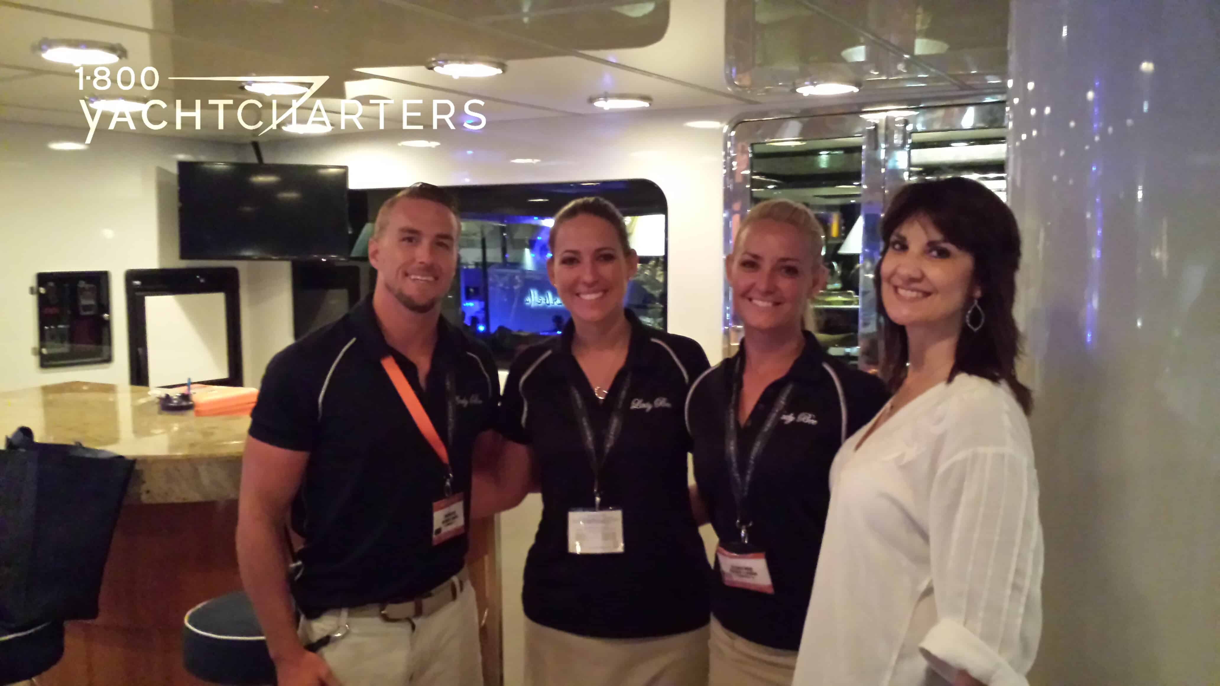 Photograph of 4 people, smiling at the camera. They are standing in the main salon of a superyacht. The three people on the left of the photo are wearing black t-shirts and name badges on lanyards. The person on the right side of the photo is wearing a white blouse and gold name badge. 