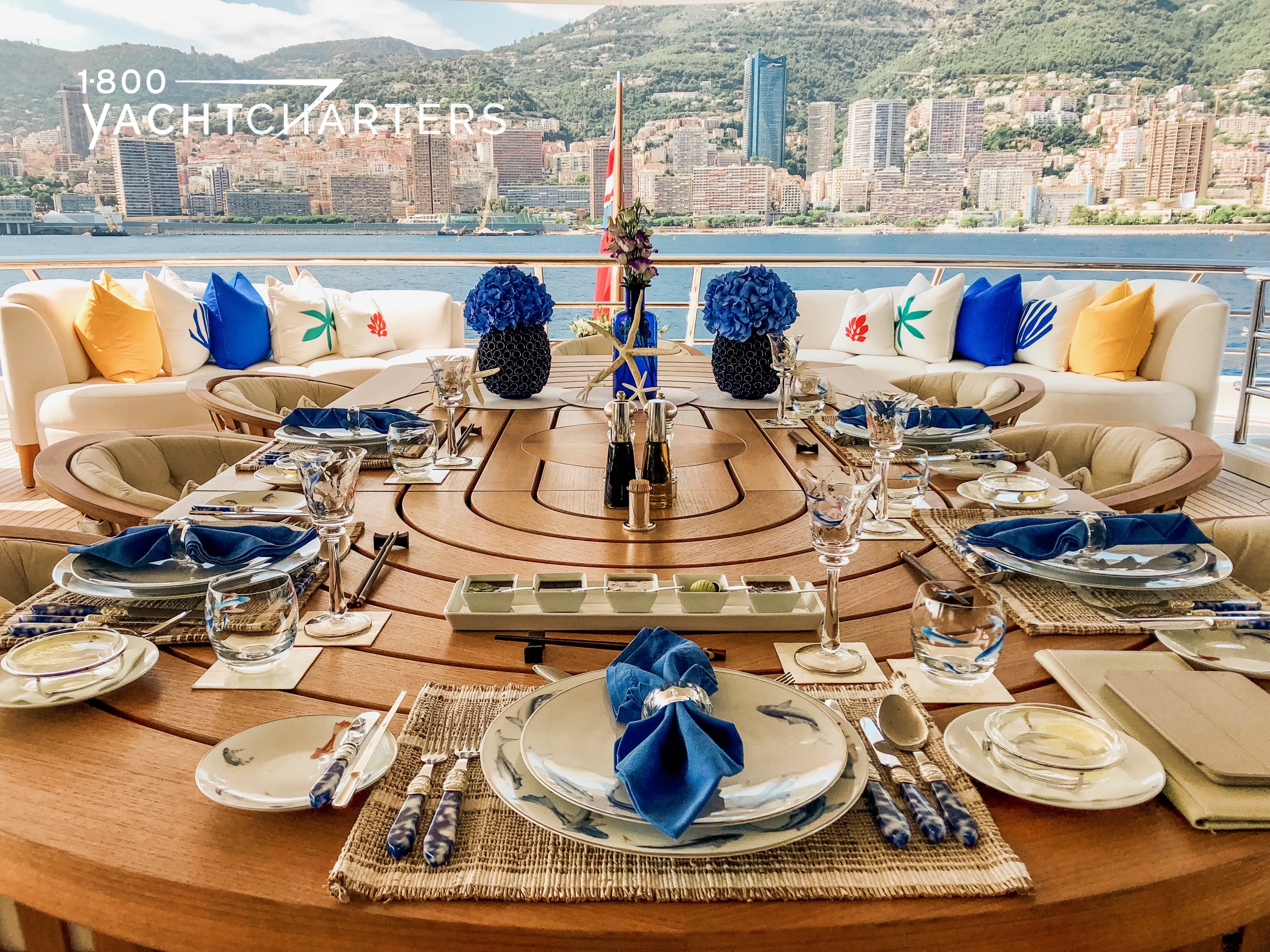 Photo taken from the back entrance of a yacht toward the back of the yacht. The yacht is facing away from a gorgeous Mediterranean cityscape. There is a table in the foreground that is elegantly set. It features blue, white, and beige. It is a large table on the deck of a yacht.