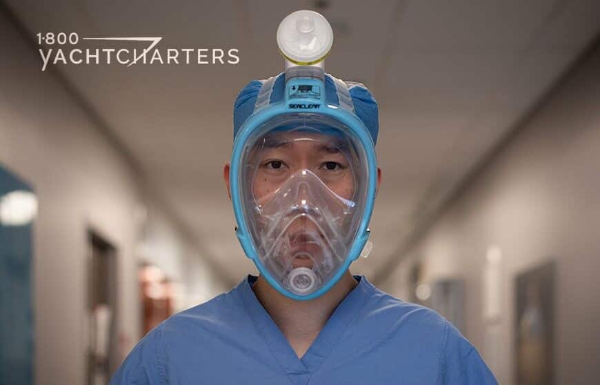 Photo of a chinese man standing in a hospital corridor. He is looking at the camera. He is wearing blue scrubs. He is wearing a light blue snorkel mask that has been retrofitted as a mask and ventilator to use by first responders for the Coronavirus Covid-19 pandemic.