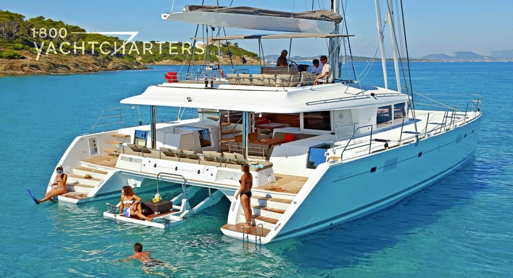Photograph of a family enjoying a charter on a catamaran sailboat at anchor. They are playing in the water and sunbathing on the deck. The water is clear and turquoise. There is an island in the background. The sailboat is all white. There are 6 people on the boat. The boat is facing the upper right side of the photograph