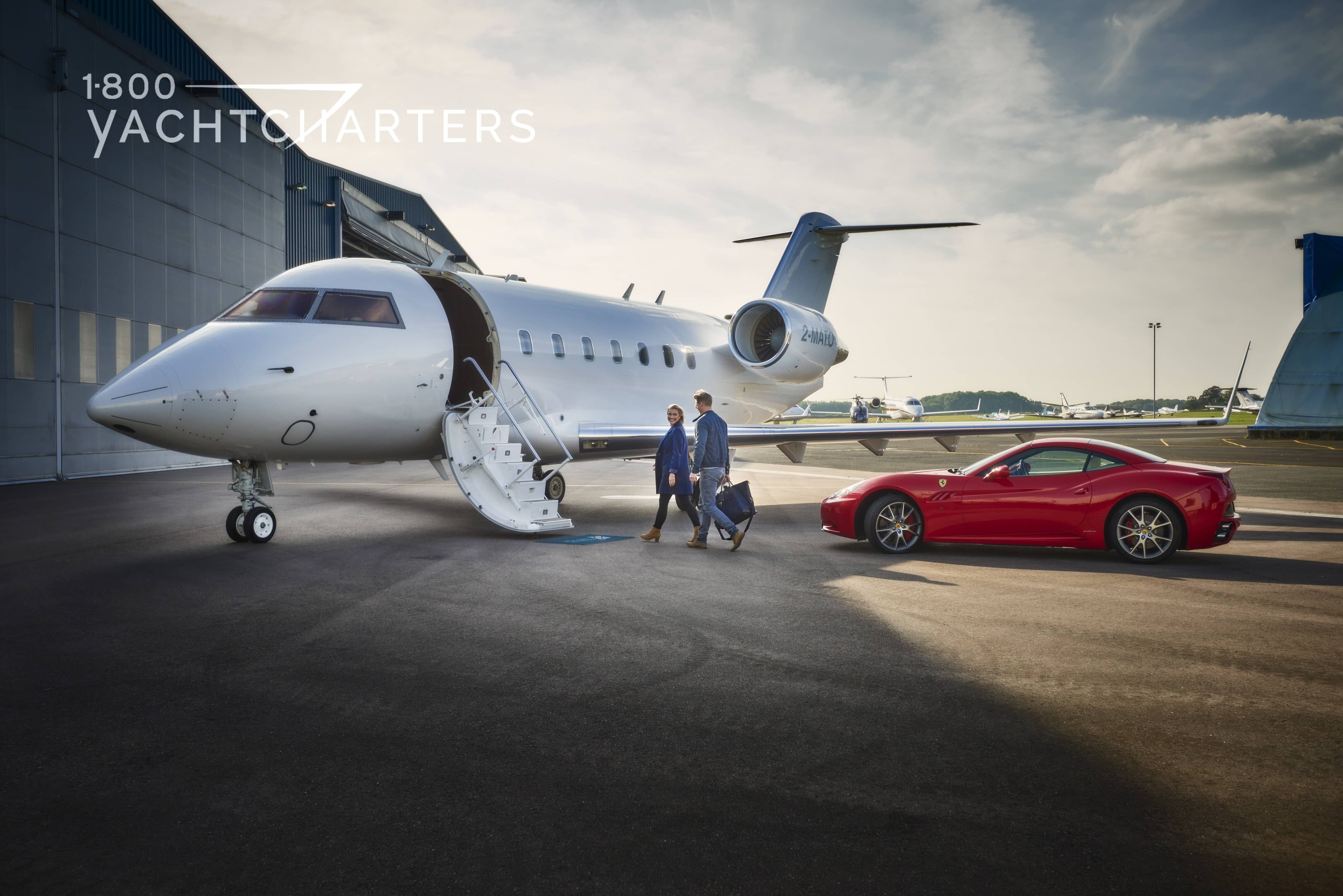 Photograph of a private jet with a red ferrari next to it. The jet is on the tarmac. The jet door is open, and the stairs are deployed. 2 men are walking up to the jet.